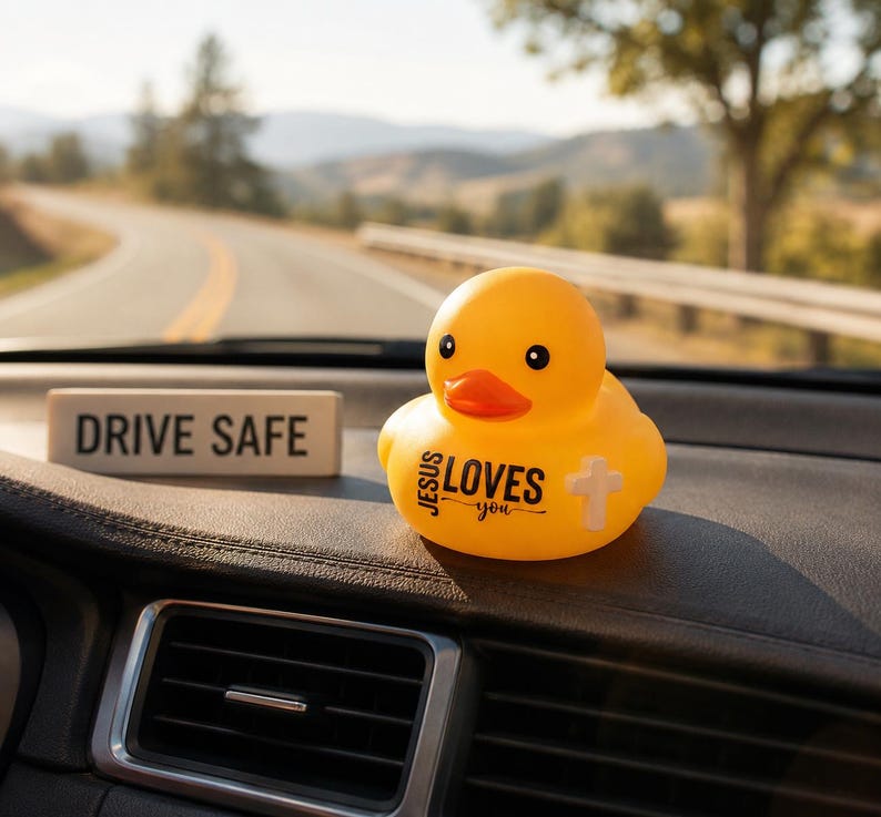 May include: A yellow rubber duck with the text "JESUS LOVES you" and a cross graphic. A small rectangular sign with "DRIVE SAFE" is also visible. The items are on a car dashboard with a scenic road in the background.