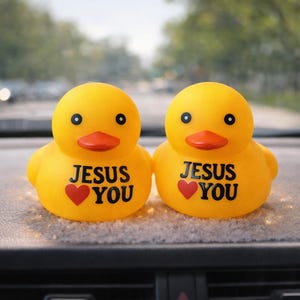 May include: Two yellow rubber duck toys with black eyes and orange beaks. Each duck has the words "JESUS YOU" printed on its chest, with a red heart below. The ducks are sitting on a car dashboard.