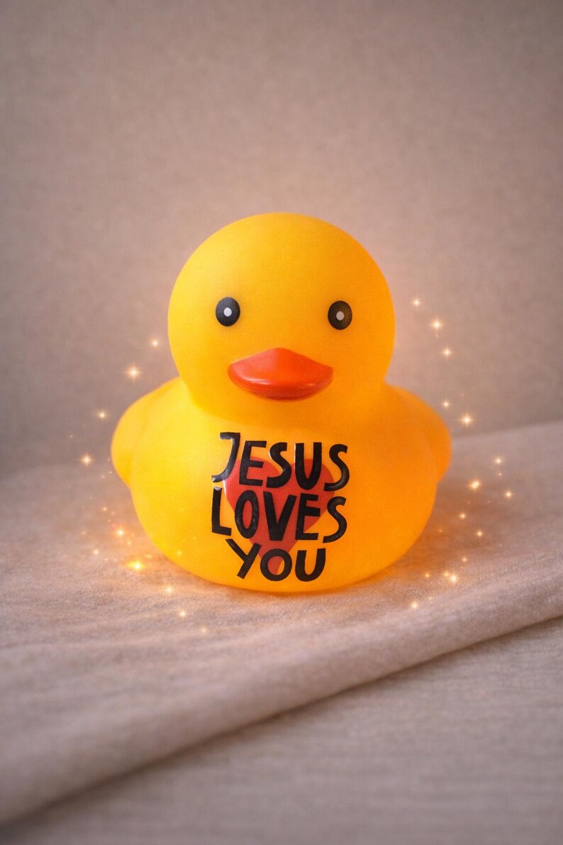 May include: A yellow rubber duck with black eyes and an orange beak. The text "JESUS LOVES YOU" is printed in black on the duck's chest. The duck is a religious-themed item.