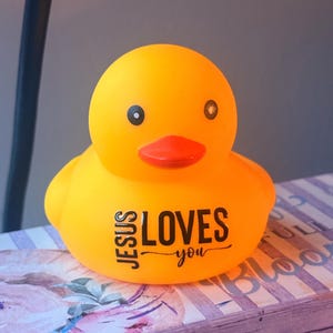 May include: A vibrant yellow rubber duck with a red beak and black eyes. The duck features the text "JESUS LOVES you" in black. It rests on a surface with floral and striped patterns.