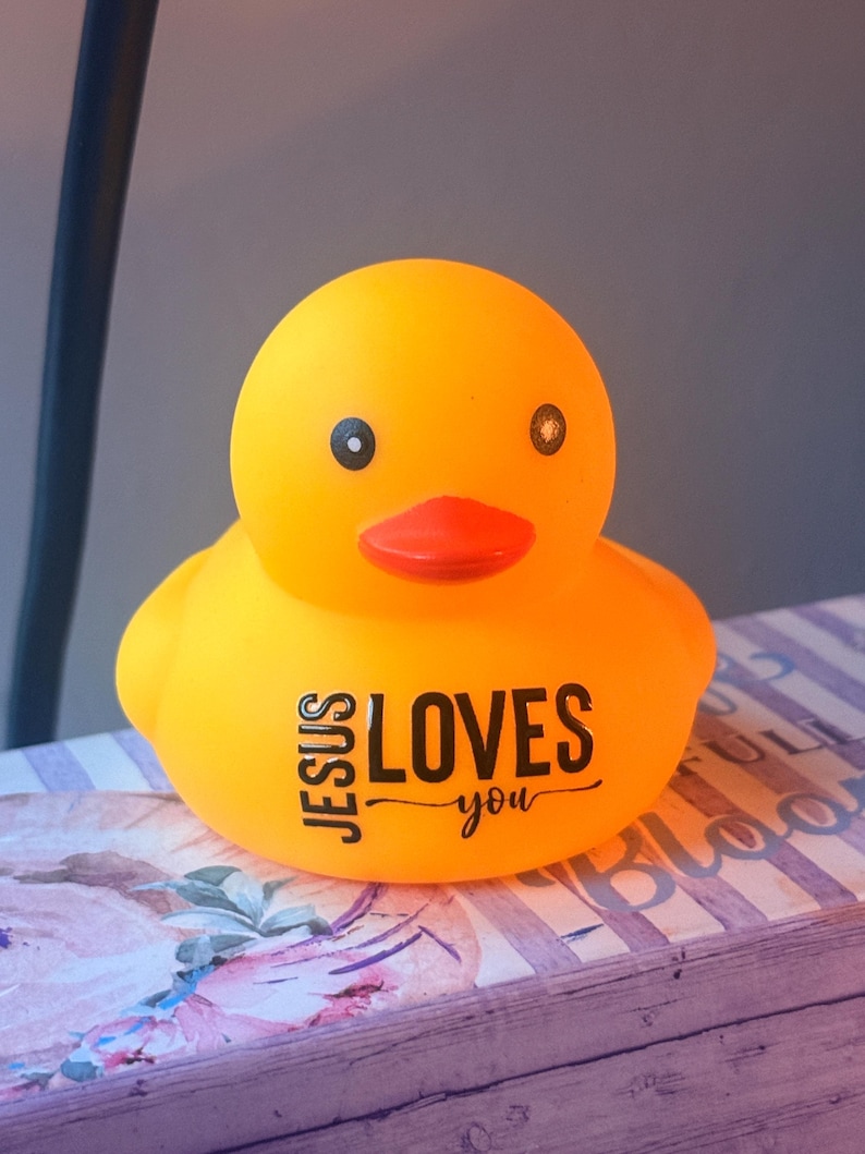 May include: A vibrant yellow rubber duck with a red beak and black eyes. The duck features the text "JESUS LOVES you" in black. It rests on a surface with floral and striped patterns.