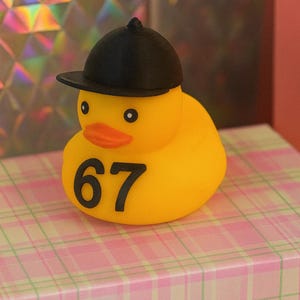 May include: A yellow rubber duck with a black baseball cap and orange beak. The number '67' is printed in black on the duck's body. The duck sits on a pink and white plaid surface.