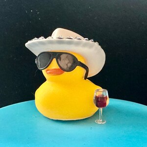 May include: A yellow rubber duck wearing a white cowboy hat with rhinestones and black sunglasses, holding a small glass of red wine. The duck is sitting on a blue surface.