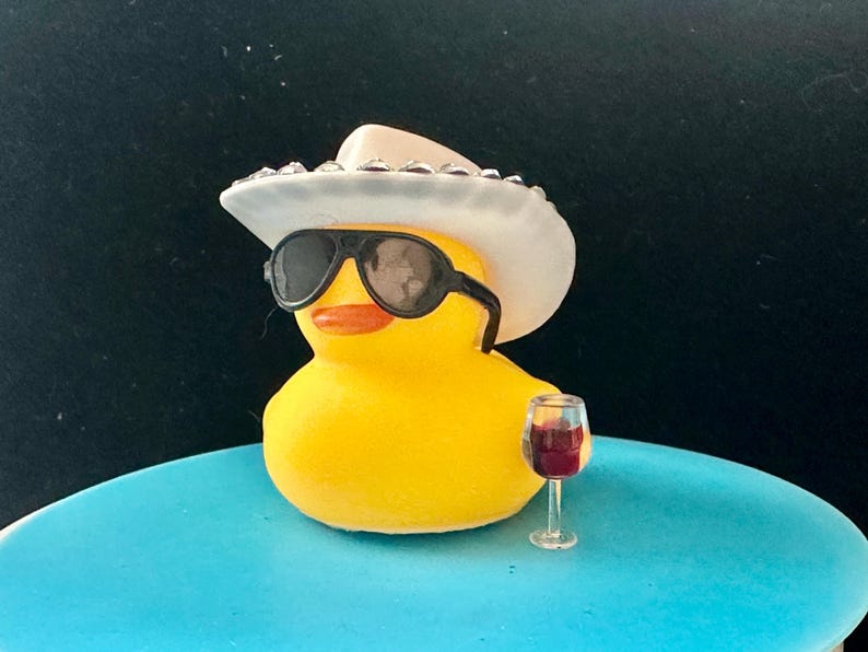 May include: A yellow rubber duck wearing a white cowboy hat with rhinestones and black sunglasses, holding a small glass of red wine. The duck is sitting on a blue surface.