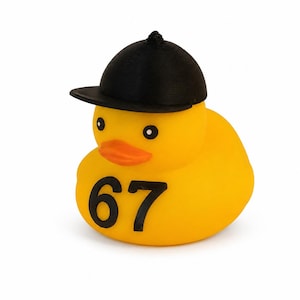 May include: A yellow rubber duck featuring a black equestrian-style helmet on its head and the black number 67 printed on its chest. The duck has a bright orange beak and two small black eyes, maintaining a classic duck shape and smooth texture.