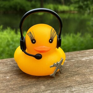 May include: A bright yellow rubber duck featuring long black eyelashes, wearing a black headset with a microphone boom, and adorned with a small silver-toned metal airplane charm attached to its side, creating a whimsical aviation-themed character.