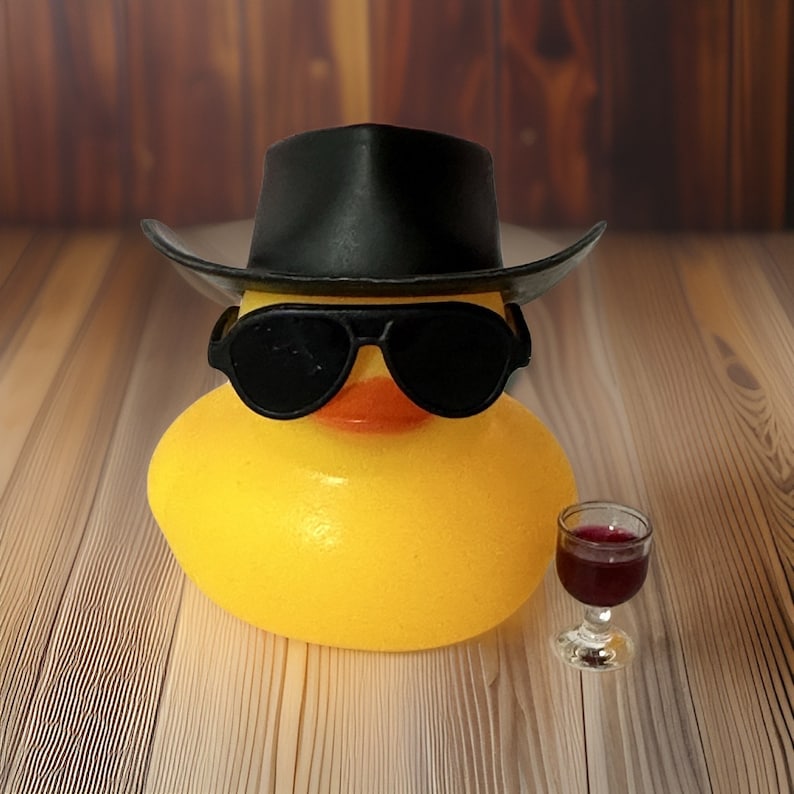 May include: A yellow rubber duck wearing a black cowboy hat and sunglasses. The duck is holding a glass of red wine.