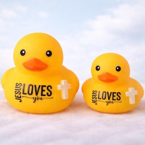May include: Two yellow rubber ducks with orange beaks and black eyes. The ducks have the text "JESUS LOVES you" printed on their bodies, along with a white cross. The ducks are on a white surface with a blue sky background.