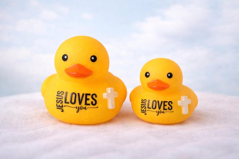 May include: Two yellow rubber ducks with orange beaks and black eyes. The ducks have the text "JESUS LOVES you" printed on their bodies, along with a white cross. The ducks are on a white surface with a blue sky background.