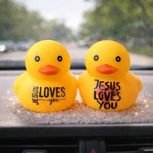 May include: Two yellow rubber ducks with orange beaks and black eyes. Each duck has the phrase "JESUS LOVES you" printed on its side in black and red. The ducks are sitting on a grey surface.