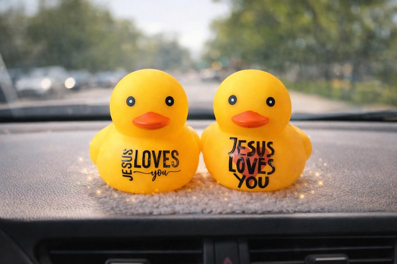 May include: Two yellow rubber ducks with orange beaks and black eyes. Each duck has the phrase "JESUS LOVES you" printed on its side in black and red. The ducks are sitting on a grey surface.