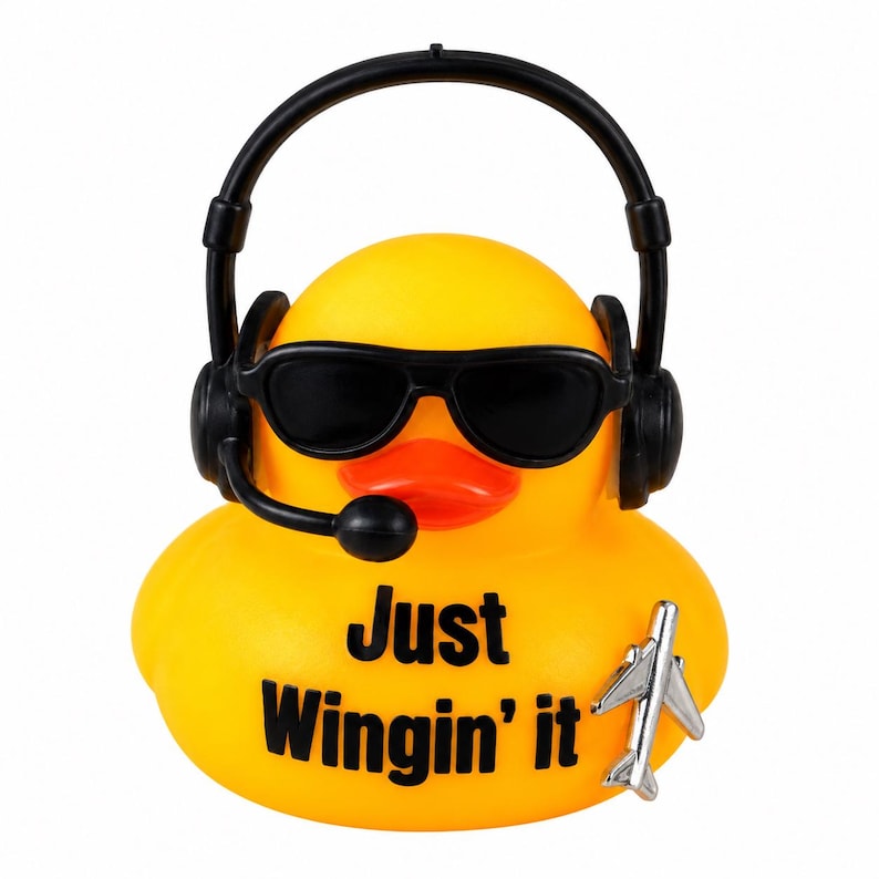 May include: A bright yellow rubber duck wearing black sunglasses and a black headset with a microphone. The duck features the black text Just Wingin' it printed on its chest and a small silver airplane figurine attached to its lower right side.
