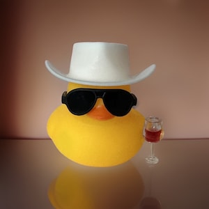 May include: A yellow rubber duck wearing a white cowboy hat and black sunglasses, holding a small glass of red wine.