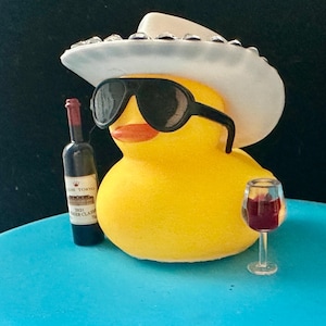 May include: A yellow rubber duck wearing a white cowboy hat with rhinestones and sunglasses, holding a bottle of red wine and a glass of red wine. The duck is sitting on a blue surface.