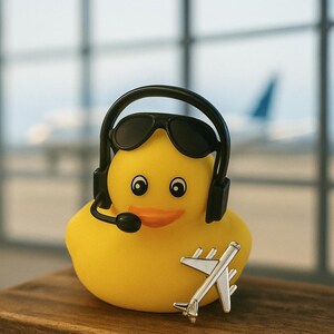 May include: A bright yellow rubber duck wearing black sunglasses and a black headset with a microphone. A small, silver-colored metal airplane figurine is positioned in front of the duck. The duck has a vibrant orange beak and black eyes.