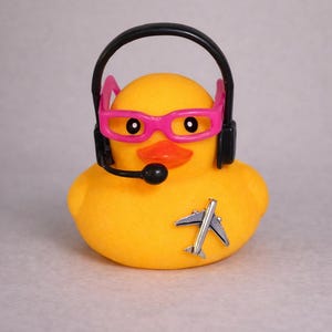May include: A yellow rubber duck wearing bright pink glasses and a black headset with a microphone. A small silver airplane pin is attached to the front of the duck. The duck has a bright orange beak and black eyes, creating a distinct character design.
