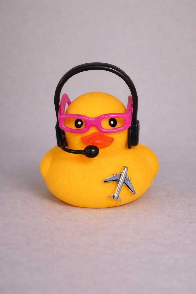 May include: A yellow rubber duck wearing bright pink glasses and a black headset with a microphone. A small silver airplane pin is attached to the front of the duck. The duck has a bright orange beak and black eyes, creating a distinct character design.