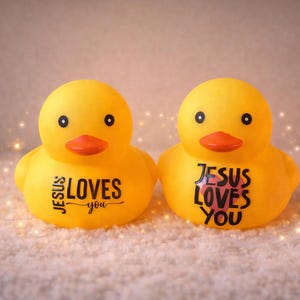 May include: Two yellow rubber duck toys with orange beaks and black eyes. Each duck has the phrase "JESUS LOVES YOU" printed on its front in black. The ducks are on a white, textured surface.
