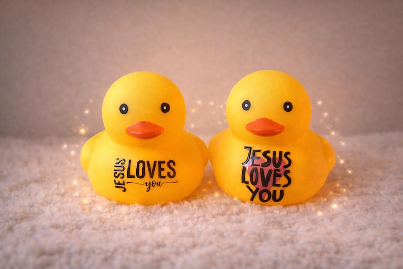 May include: Two yellow rubber duck toys with orange beaks and black eyes. Each duck has the phrase "JESUS LOVES YOU" printed on its front in black. The ducks are on a white, textured surface.