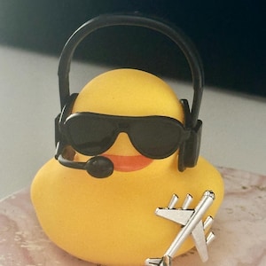 May include: A yellow rubber duck wearing black sunglasses and a black headset with a microphone. A small silver-colored airplane charm is attached to the front of the duck. The duck has an orange beak and a smooth, rounded body shape.