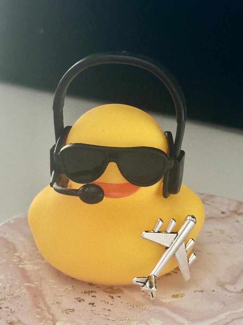 May include: A yellow rubber duck wearing black sunglasses and a black headset with a microphone. A small silver-colored airplane charm is attached to the front of the duck. The duck has an orange beak and a smooth, rounded body shape.
