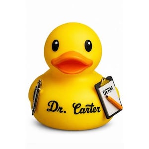 Puede incluir: Un pato de goma amarillo brillante con el nombre Dr. Carter impreso en negro en el pecho. El pato sostiene un bolígrafo plateado en un lado y un portapapeles negro con el texto DERM y una ilustración de un lápiz naranja en el otro.