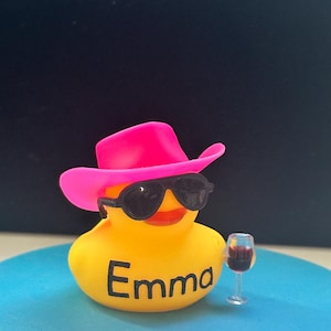 May include: A yellow rubber duck wearing a pink cowboy hat and sunglasses, holding a glass of red wine. The duck is sitting on a blue surface and has the name "Emma" written on its chest.