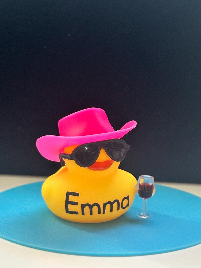 May include: A yellow rubber duck wearing a pink cowboy hat and sunglasses, holding a glass of red wine. The duck is sitting on a blue surface and has the name "Emma" written on its chest.