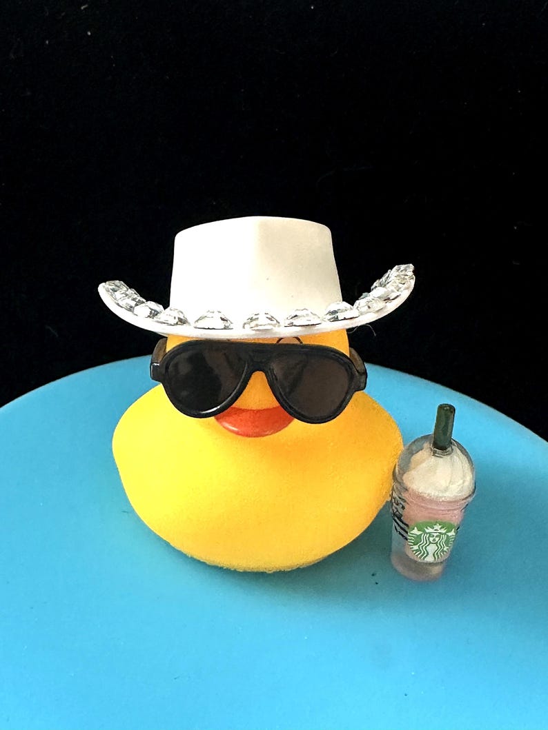 Może przedstawiać: Ż&oacute;łta gumowa kaczka w okularach przeciwsłonecznych i białym kowbojskim kapeluszu z cyrkoniami. Kaczka trzyma r&oacute;żowo-zielony Frappuccino Starbucks.