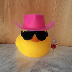 May include: A yellow rubber duck wearing a pink cowboy hat and sunglasses, holding a small glass of red wine.