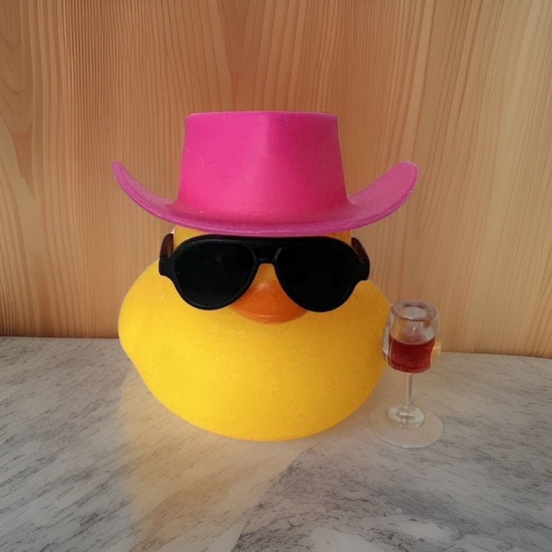 May include: A yellow rubber duck wearing a pink cowboy hat and sunglasses, holding a small glass of red wine.