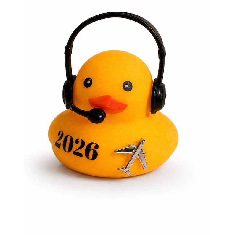 May include: A yellow rubber duck wearing black headphones with a microphone. The duck features the black text 2026 printed on its side and a small silver-colored airplane emblem attached to its body, creating a distinct and recognizable figure.