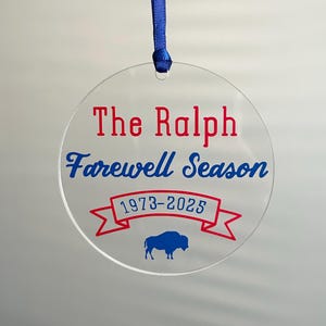 May include: A clear, circular ornament with the text "The Ralph Farewell Season 1973-2025" in red and blue. A blue buffalo silhouette is at the bottom. A blue ribbon is attached for hanging.