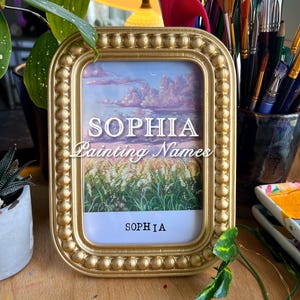 May include: A gold-colored picture frame with a painting of a field of grass and a cloudy sky. The name "Sophia" is written in white letters on the frame and the painting.