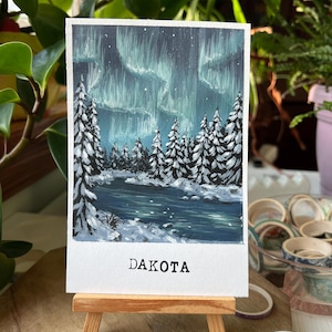 May include: A small painting on a wooden easel depicts a winter landscape with a river and snow-covered trees under a vibrant aurora borealis. The word "DAKOTA" is printed at the bottom of the painting.