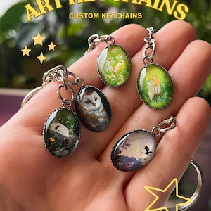 May include: Oval art keychains with various miniature paintings, including an owl, a field of daisies, and a dragon silhouette. The keychains are silver-toned and attached to key rings. Text on the image reads "ART KEYCHAINS" and "CUSTOM KEYCHAINS".