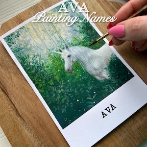 May include: A painting of a white unicorn in a forest setting, with green foliage and a soft, light background. The artwork is on a white rectangular card, with the name "AVA Painting Names" and "AVA" printed on it. The painting is on a wooden surface.