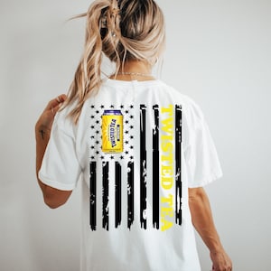 May include: White t-shirt with a black and white American flag design. The flag has a yellow "Twisted Tea" text and a yellow can of Twisted Tea with a purple label.