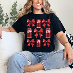 May include: A black t-shirt featuring a red and white Dr Pepper can design. The cans are surrounded by red bows. The shirt is being worn by a person with long blonde hair and blue jeans.