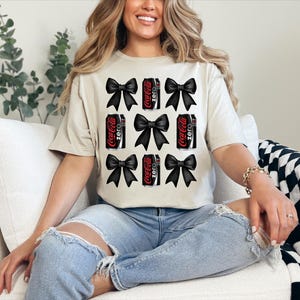 Könnte beinhalten: Ein weißes T-Shirt mit einem schwarzen und roten Coca-Cola Zero-Design mit schwarzen Schleifen. Das Shirt hat kurze Ärmel und einen Rundhalsausschnitt.