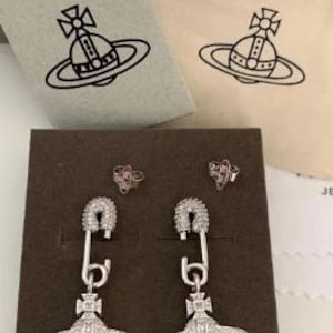 K&ouml;nnte beinhalten: Silberfarbene Ohrringe im Sicherheitsnadel-Design mit einem planeten&auml;hnlichen Anh&auml;nger. Pr&auml;sentiert auf einer braunen Schachtel, zusammen mit zwei kleinen Ohrsteckern und zwei Beuteln mit Logo.
