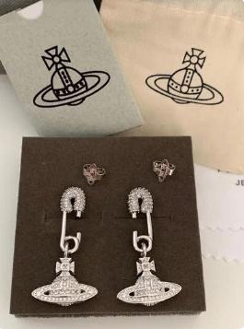 K&ouml;nnte beinhalten: Silberfarbene Ohrringe im Sicherheitsnadel-Design mit einem planeten&auml;hnlichen Anh&auml;nger. Pr&auml;sentiert auf einer braunen Schachtel, zusammen mit zwei kleinen Ohrsteckern und zwei Beuteln mit Logo.