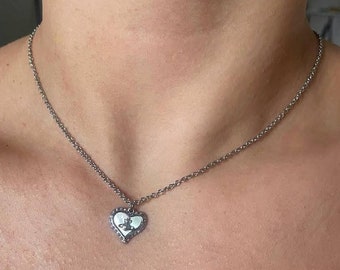 Op de afbeelding: Zilverkleurige ketting met een hartvormige hanger. De hanger heeft een witte kern met een klein ontwerp en is omgeven door een rand van kleine, donkere stenen. De ketting is een delicaat, geschakeld ontwerp.