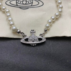 Op de afbeelding: Een zilveren ketting met een parelketting en een hanger in de vorm van een bol bedekt met kristallen. De hanger heeft een klein kruis aan de bovenkant.