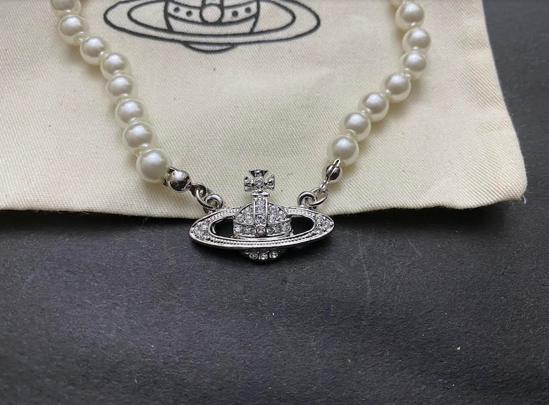 Op de afbeelding: Een zilveren ketting met een parelketting en een hanger in de vorm van een bol bedekt met kristallen. De hanger heeft een klein kruis aan de bovenkant.