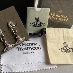 K&ouml;nnte beinhalten: Silberfarbene Ohrringe im Sicherheitsnadel-Design, verziert mit glitzernden Kristallen. Die Ohrringe werden zusammen mit Markenverpackungen gezeigt, darunter eine Schachtel, ein Beutel und ein Reinigungstuch, alle mit dem Vivienne Westwood-Logo.