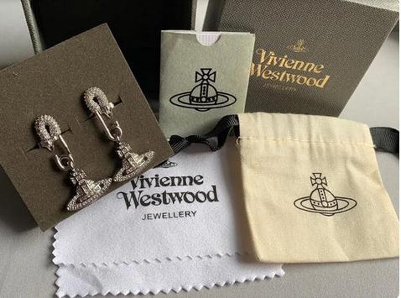 K&ouml;nnte beinhalten: Silberfarbene Ohrringe im Sicherheitsnadel-Design, verziert mit glitzernden Kristallen. Die Ohrringe werden zusammen mit Markenverpackungen gezeigt, darunter eine Schachtel, ein Beutel und ein Reinigungstuch, alle mit dem Vivienne Westwood-Logo.