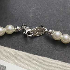 Op de afbeelding: Een zilveren ketting met een zilveren sluiting en witte parels. De sluiting heeft de tekst "Vivienne Westwood" gegraveerd.