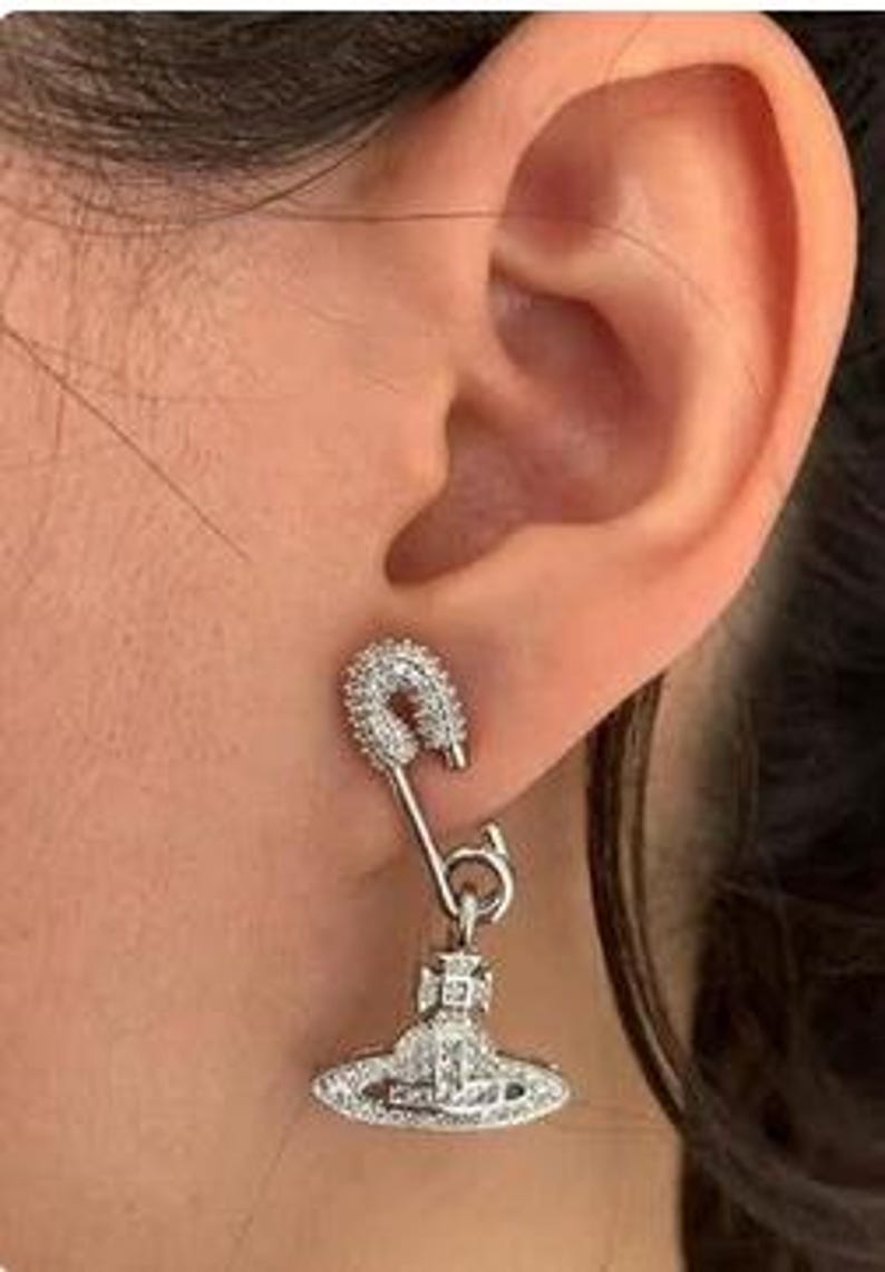 K&ouml;nnte beinhalten: Silberner Ohrring mit Sicherheitsnadel-Design und einem h&auml;ngenden Anh&auml;nger. Der Anh&auml;nger hat eine stilisierte Orb-Form, ebenfalls silberfarben, und beide Teile sind mit kleinen, klaren Kristallen besetzt.