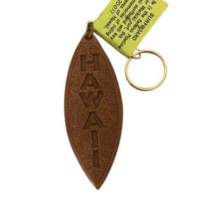 Może przedstawiać: Brelok w kształcie brązowej deski surfingowej z wytłoczonym napisem "HAWAII". Dołączona żółta etykieta głosi "SURFBOARD" i wspomina Pipeline i Waikiki na Hawajach. Dołączony jest złoty brelok.